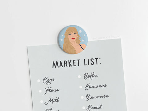 A market list on white paper is held by the TS Midnights Button Magnet from Onderkast Studio, a round magnet featuring an illustrated woman with long blonde hair, a black strap, and celestial blue background with white stars.