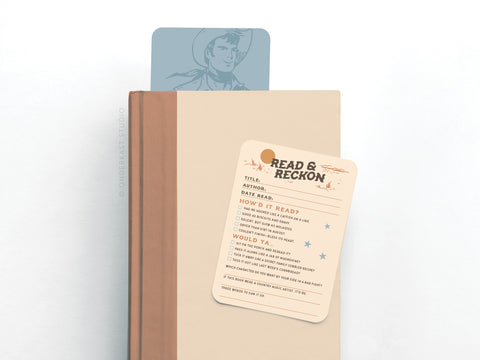 A beige hardcover book with a blue bookmark features a cowboy illustration and is topped with an Onderkast Studio Read & Reckon Book Review Card Set, which includes sections for title, author, and reading notes.
