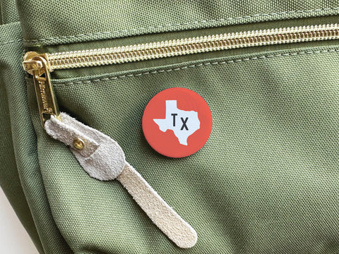 The Red TX Pinback Button by Onderkast Studio, featuring a white Texas outline, is pinned to a light green fabric bag with a gold zipper and tan suede pull.