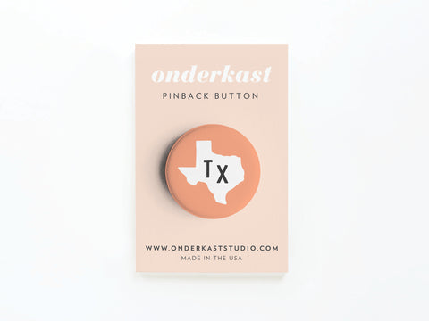An Orange TX Pinback Button from Onderkast Studio shows a white Texas silhouette with black TX letters on orange, packaged on a beige Onderkast PINBACK BUTTON card with website and MADE IN THE USA text below, highlighting quality craftsmanship.