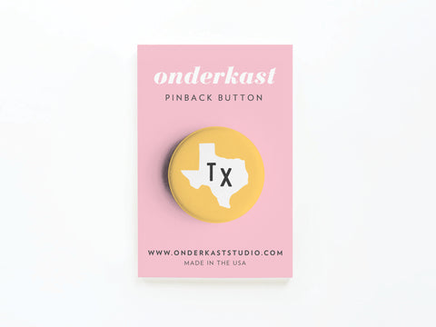 The Yellow TX Pinback Button by Onderkast Studio features a white Texas state outline and Tx text on yellow, displayed on a pink card labeled “onderkast pinback button” with onderkaststudio.com and “Made in the USA.”.