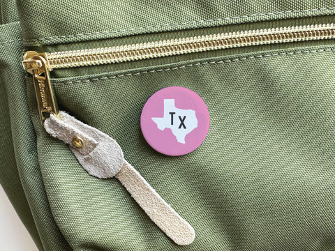The Pink TX Pinback Button by Onderkast Studio features a white Texas state outline and Tx text, showcasing Texas Pride as it’s pinned to a green bag with a gold zipper and beige suede pull.