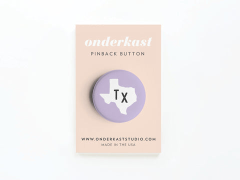 The Purple TX Pinback Button by Onderkast Studio features a white Texas silhouette with black Tx on a lavender background, attached to a branded beige card—ideal for displaying your state pride.