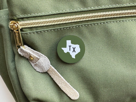 The Dark Green TX Pinback Button by Onderkast Studio, featuring a white Texas outline and TX in the center, is attached to a green bag near a zippered pocket—a stylish way to show your state pride.