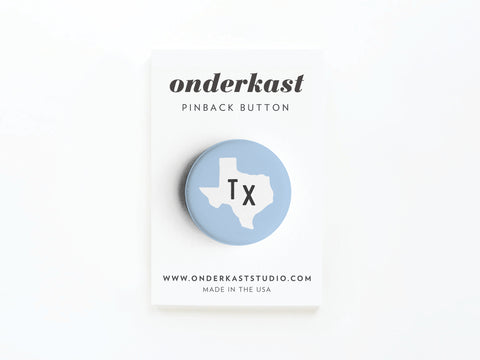 The Light Blue TX Pinback Button by Onderkast Studio features a white Texas silhouette and “Tx” on blue, displayed on a branded card—perfect for showing your Texas pride.