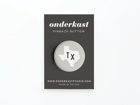The Gray TX Pinback Button by Onderkast Studio features a white Texas outline with black TX lettering, vibrant colors, and MADE IN THE USA, all displayed on a branded card with website details.