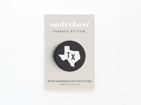 The Black TX Pinback Button by Onderkast Studio features a bold TX and white Texas outline, comes attached to a card with onderkast PINBACK BUTTON, www.onderkaststudio.com, and is printed in the USA.