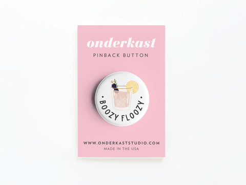 The Boozy Floozy Cocktail Pinback Button by Onderkast Studio features a playful illustrated cocktail with lemon and herbs, labeled BOOZY FLOOZY, and is displayed on a pink Onderkast Studio card.