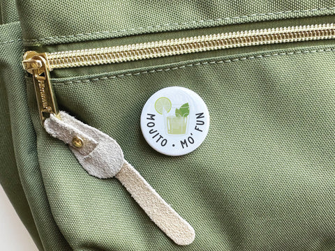 The Onderkast Studio Mojito Mo’ Fun Pinback Button, featuring a mojito with lime and mint, is shown on a green bag with a gold zipper and beige suede pull. Printed in the USA, the button reads MOJITO MO FUN.