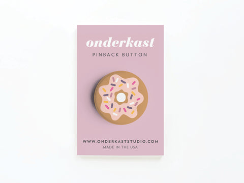 The Sprinkle Donut Pinback Button by Onderkast Studio, featuring pink and white sprinkles, comes on a pink card labeled Onderkast Pinback Button with website and Made in the USA details—ideal for donut lovers!.