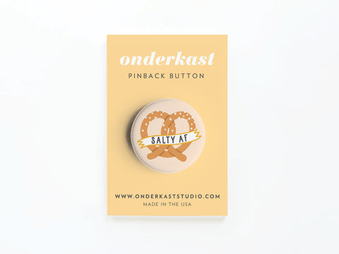 The Salty AF Pretzel Pinback Button by Onderkast Studio features a pretzel illustration and banner, ideal for snack lovers. Displayed on a yellow Onderkast card with website and MADE IN THE USA text at the bottom.