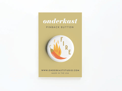 The Fire Chili Pinback Button by Onderkast Studio features a bold chili pepper with orange flames and the word FIRE. It comes on a tan card labeled ONDERKAST PINBACK BUTTON and ONDERKASTSTUDIO.COM, with vibrant, high-quality printing.