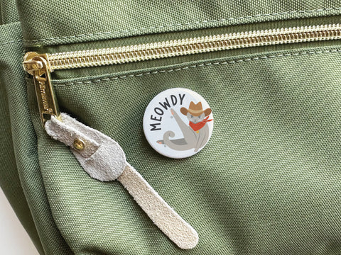 The Meowdy Pinback Button by Onderkast Studio, featuring a cat in a cowboy hat and red bandana with “MEOWDY” above, decorates a green fabric bag with visible metallic zipper and suede pull tab—perfect for cat lovers.