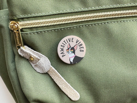The Onderkast Studio Pawsitive Vibe Dog Pinback Button featuring a happy pup is attached to a light green backpack with a gold zipper, adding style and charm.