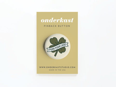 The Push Your Luck Clover Pinback Button by Onderkast Studio features a shamrock and PUSH YOUR LUCK text, displayed on a beige Onderkast card. Website and Made in the USA are printed at the bottom.