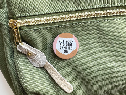 The Onderkast Studio Put On Your Big Girl Panties Pinback Button makes a bold statement on a light green fabric bag with a gold zipper and tan suede pull, empowering women with its message at every glance.