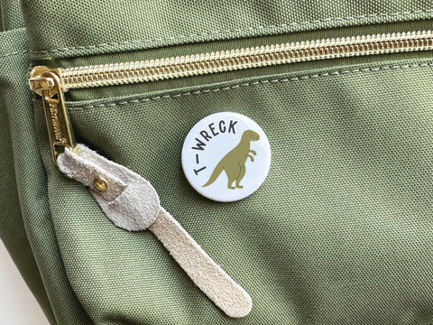 A green fabric bag with a gold zipper and suede pull. Attached is a discontinued Onderkast Studio “T-wreck” pinback button, vibrantly printed with a cartoon dinosaur and text.