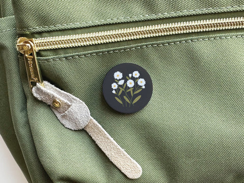 The Onderkast Studio Daisies Pinback Button, a round black button with a white daisy design, is attached to the front pocket of a light green backpack featuring a gold zipper and beige suede pull.