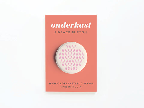 The YASS Pink Pinback Button by Onderkast Studio features rows of pink As with a few Y and S, printed vibrantly and mounted on a coral card labeled onderkast pinback button and www.onderkaststudio.com. Made in the USA. Discontinued.