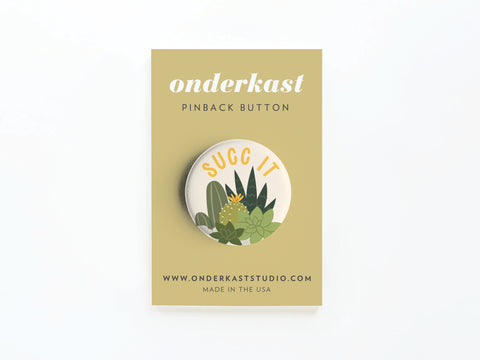 The Succ It Succulents Pinback Button by Onderkast Studio features a vibrant succulent design and SUCC IT text, displayed on a beige card—perfect for plant lovers. www.onderkaststudio.com Made in the USA.