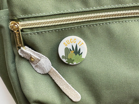The Succ It Succulents Pinback Button by Onderkast Studio, with “SUGG IT” above cute succulent art, adds charm to any green backpack—ideal for plant lovers.