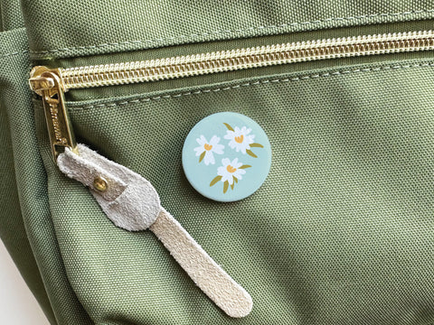 A light green fabric backpack with a gold zipper and beige suede pull features the Onderkast Studio White Flowers Pinback Button, which showcases a charming mint-green design with white and yellow flowers and green leaves.