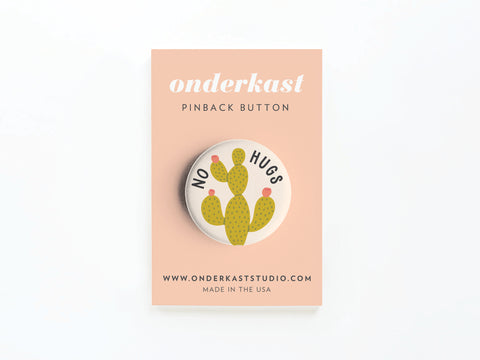 The No Hugs Cactus Pinback Button by Onderkast Studio features a green cactus and “NO HUGS” text—great for introverts. Displayed on a peach card with onderkast PINBACK BUTTON, website, and MADE IN THE USA label.