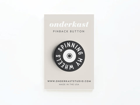 The Spinning My Wheels Pinback Button – DISCONTINUED by Onderkast Studio features white text on black, attached to an Onderkast PINBACK BUTTON display card with vibrant print and MADE IN THE USA at the bottom.