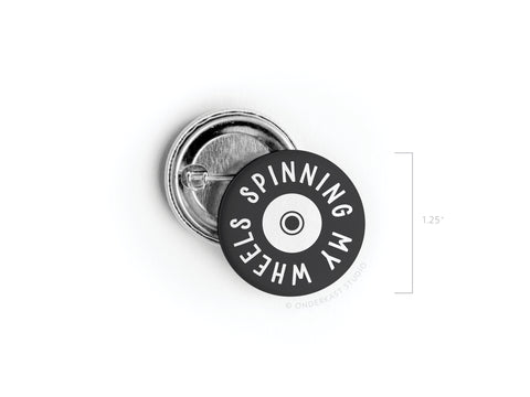 The Onderkast Studio Spinning My Wheels Pinback Button (DISCONTINUED) is a 1.25 round black and white button with SPINNING MY WHEELS encircling a small wheel illustration, shown front and back.