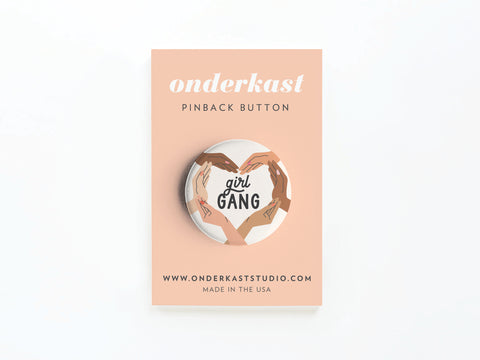 The Girl Gang Pinback Button by Onderkast Studio features illustrated hands forming a heart around girl GANG on a peach card, celebrating female friendship. Card includes the brand name, product details, website, and MADE IN THE USA.
