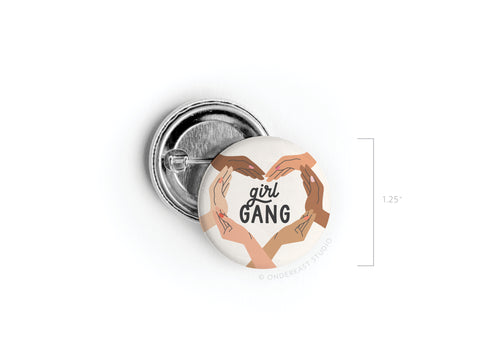 The Girl Gang Pinback Button by Onderkast Studio features illustrated diverse hands forming a heart around Girl Gang on a white background. Celebrate female friendship with this 1.25 round button.