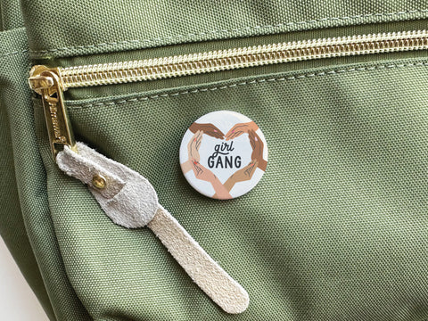The Girl Gang Pinback Button by Onderkast Studio, shown on a green fabric bag beside a gold zipper and suede pull, features girl GANG and four hands of varied skin tones forming a square to celebrate female friendship.