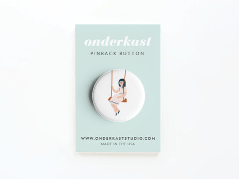 The Girl on a Swing Pinback Button by Onderkast Studio features a nostalgic illustration, displayed on a pastel green backing card labeled Onderkast pinback button with the website and Made in the USA at the bottom. High-quality and long-lasting.