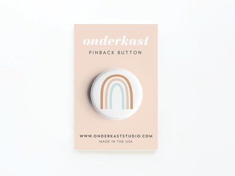 The Rainbow Pinback Button by Onderkast Studio features a minimalist pastel rainbow design on a light peach card, reading PINBACK BUTTON and www.onderkaststudio.com, celebrating inclusivity. Made in the USA.