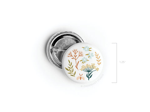 The Soft Florals Pinback Button by Onderkast Studio is a 1.25-inch round button with an illustrated wildflower design in earthy tones on white, shown beside its open back.