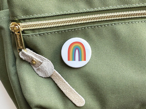 The Onderkast Studio Pride Rainbow Pinback Button, featuring a vibrant rainbow design symbolizing LGBTQ+ equality, is attached to a light green fabric bag with a gold zipper and beige suede pull.