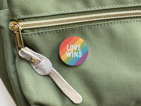 A round Love Wins Pinback Button by onderkast studio, featuring rainbow stripes and LOVE WINS text, is attached to a light green bag pocket—proudly symbolizing allyship and support for LGBTQ+ rights.