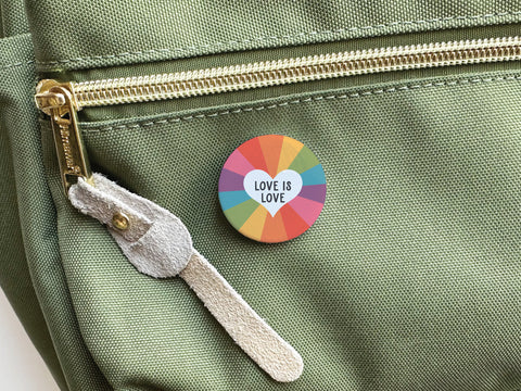 A round Love Is Love Pinback Button by onderkast studio, featuring a rainbow pattern and the phrase LOVE IS LOVE, is attached to a light green bag with a gold zipper and beige suede pull, celebrating love as universal.
