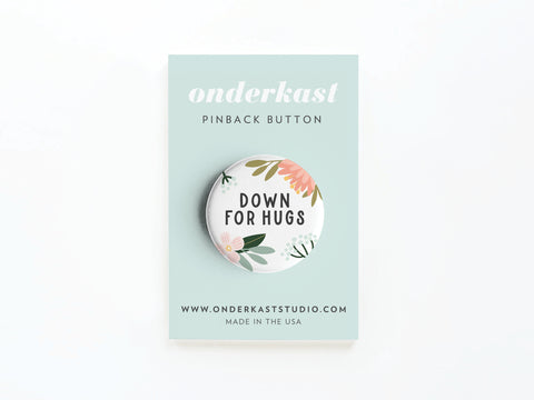 The Down For Hugs Pinback Button by Onderkast Studio features vibrant floral designs and the phrase DOWN FOR HUGS on a round button, displayed on a pastel card with branding and MADE IN THE USA printed on a light blue background.