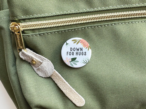 A colorful Onderkast Studio Down For Hugs Pinback Button with floral motifs is pinned to the pocket of a light green fabric bag featuring a beige suede zipper pull.