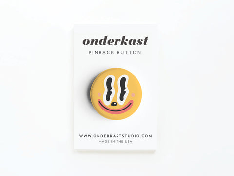 A yellow Trippy Happy Face Pinback Button from Onderkast Studio is attached to a white card labeled PINBACK BUTTON, featuring onderkaststudio.com and MADE IN THE USA.