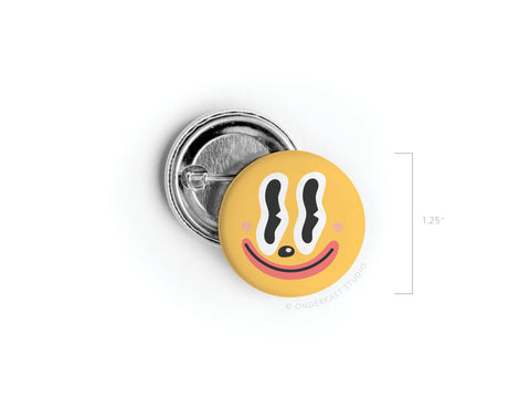 The Trippy Happy Face Pinback Button by Onderkast Studio is a 1.25-inch round, yellow button featuring a playful, wavy-eyed happy face with pink cheeks and a curved red smile, shown on a white background.