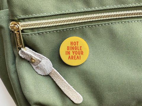 The Onderkast Studio Hot Single in Your Area! Pinback Button features bold red text on yellow, attached to a green bag with a gold zipper and tan suede pull—making it an ideal quirky online dating accessory.