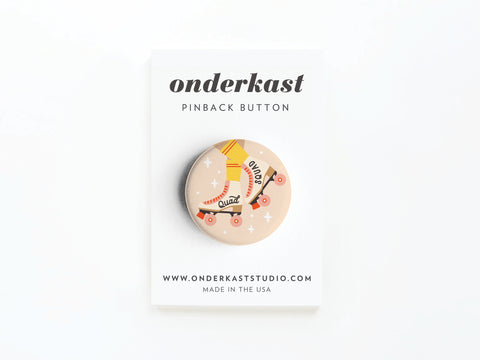 The Quad Squad Roller Skates Pinback Button by Onderkast Studio features roller skates and yellow-striped socks on a light background, attached to a labeled card. Website and MADE IN THE USA printed below. Great for any Quad Squad fan!.