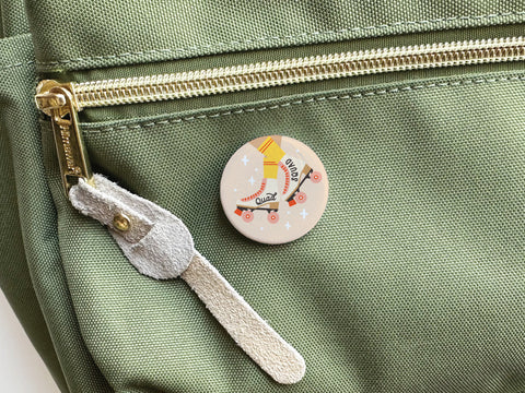 The Quad Squad Roller Skates Pinback Button by Onderkast Studio, featuring a retro skate and Old School text, is attached to the front pocket of a light green canvas bag with a gold zipper and suede pull.