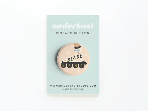The Blade Gang Rollerblades Pinback Button by Onderkast Studio comes on a light blue card with the brand’s logo, onderkaststudio.com, and Made in the USA. This fun skater accessory features a roller skate, Blade Gang text, and colorful accents.