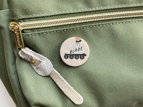 The Blade Gang Rollerblades Pinback Button by Onderkast Studio is attached to the front pocket of a light green backpack with a gold zipper and beige suede pull—a must-have skater accessory for rollerblading fans.