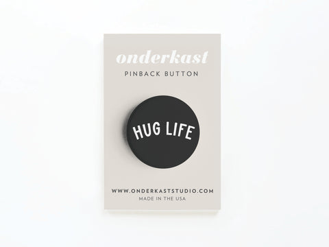 The Hug Life Pinback Button by Onderkast Studio features black with white lettering, displayed on a beige card labeled Onderkast Pinback Button with the website and Made in the USA below.