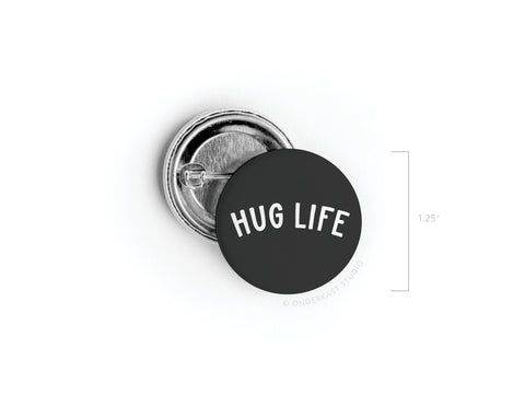 The Hug Life Pinback Button by Onderkast Studio is a 1.25-inch round button with white, curved uppercase letters, shown from both front and back angles.