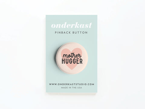 The Mother Hugger Pinback Button by Onderkast Studio features a pink heart and text, displayed on a light blue card labeled onderkast PINBACK BUTTON with onderkaststudio.com—perfect for Mother’s Day.